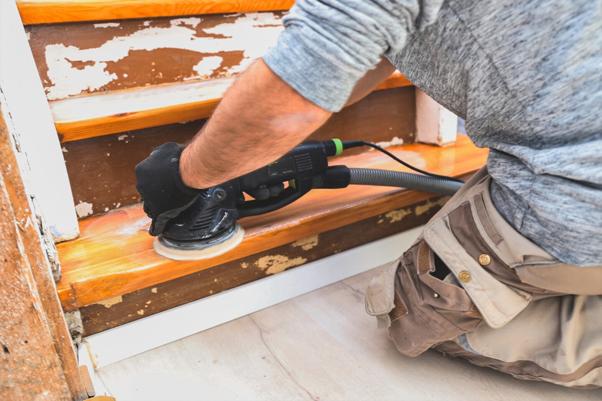 Entfernung des vorhandenen Belages bei der Treppe Ein Mann entfernt den aktuellen Belag von einer Holztreppe mithilfe eines Schwingschleifers.