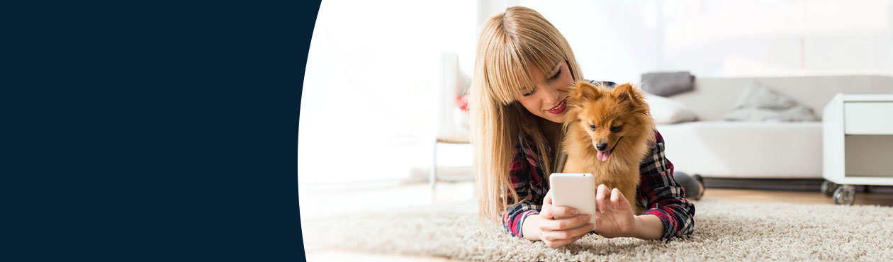 Frau mit Hund liegt auf einem Teppich und schaut in ein Smartphome