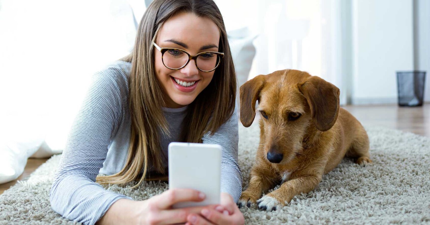 Frau mit Hund liegt auf einem Teppich und schaut in ein Smartphome