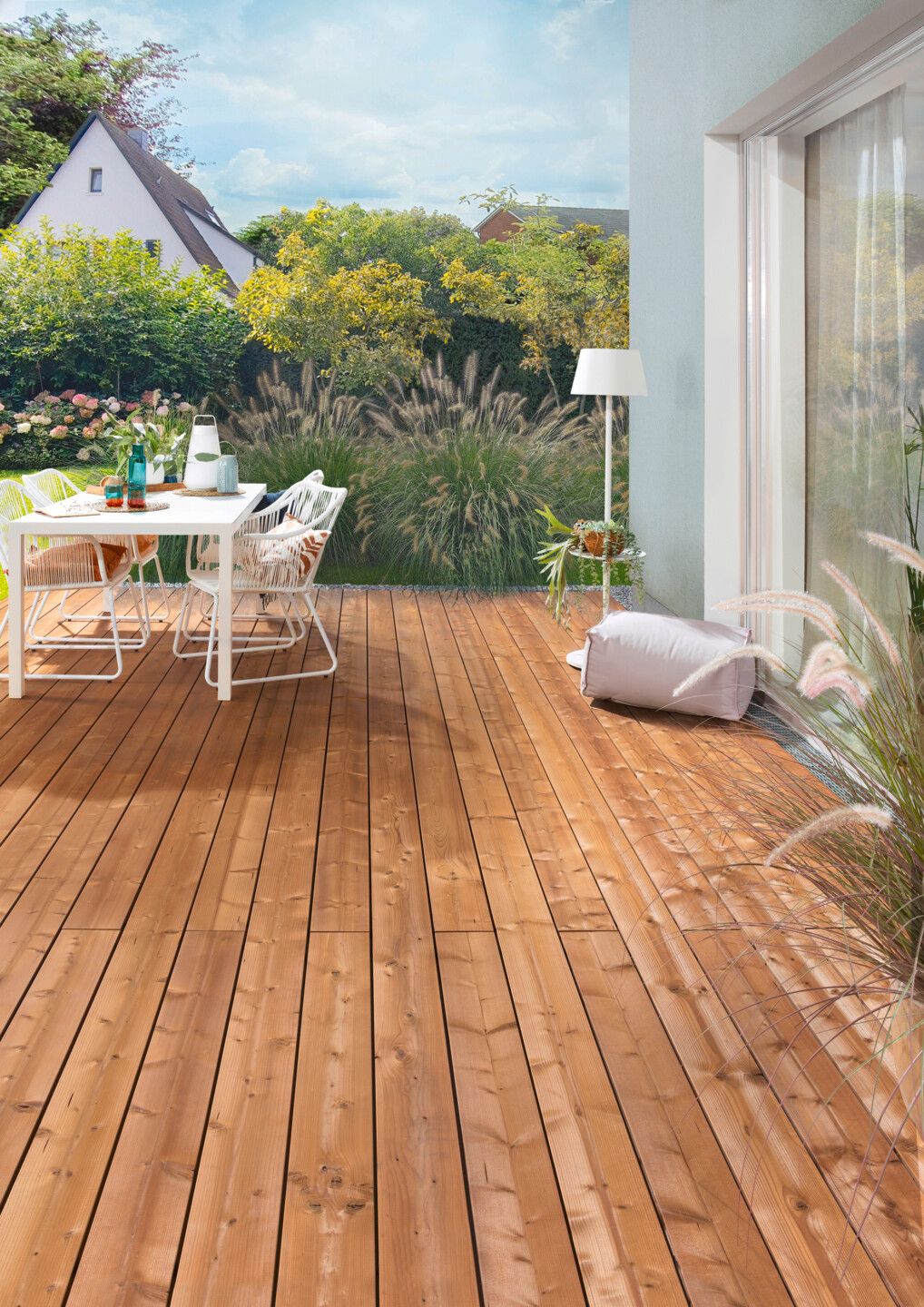 Terrasse am Haus aus Holzdielen mit moderner weißer Tischgarnitur und im Hintergrund ein modern gestalteter Garten mit Gräsern
