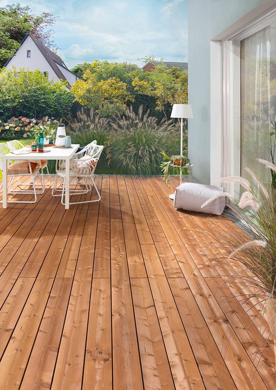 Terrasse am Haus aus Holzdielen mit moderner weißer Tischgarnitur und im Hintergrund ein modern gestalteter Garten mit Gräsern