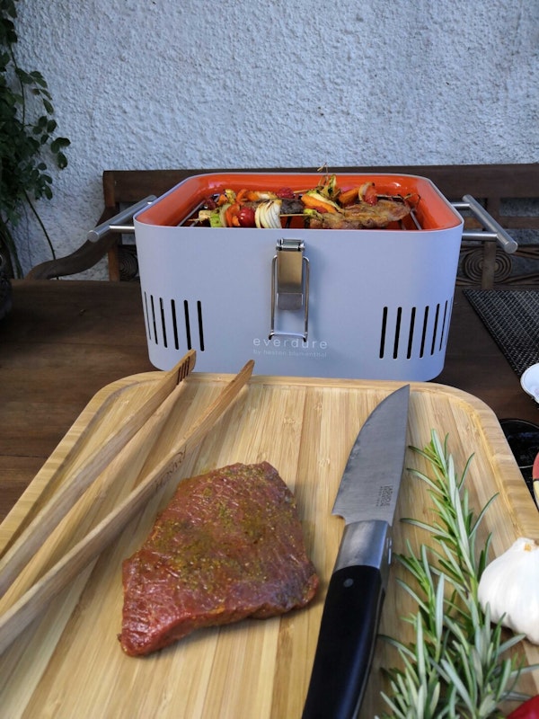 Das praktische Schneidebrett aus Bambus ist robust und leicht zu reinigen Auf dem Schneidebrett vor dem Grill liegt das nächste Stück Fleisch bereit, der Grillrost ist noch voll