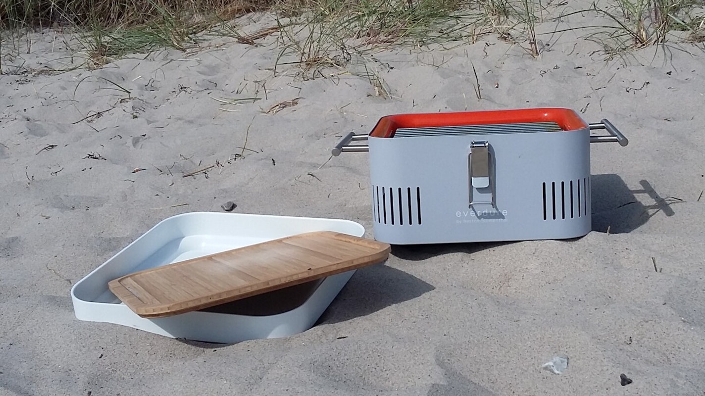 Der perfekte Urlaubsbegleiter - Everdure Cube Cube Grill steht vor einer Düne am Strand, die weiße Aufbewahrungsbox und das Schneidebrett aus Bambus liegen daneben