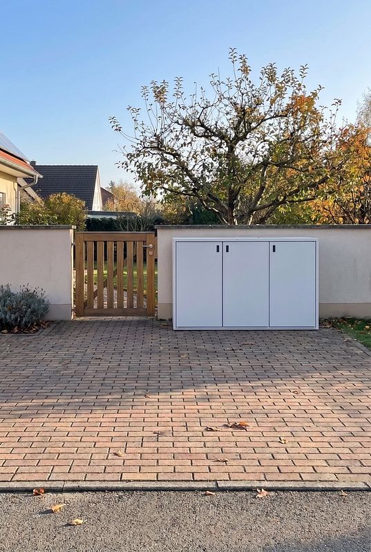Vor einer hellen Gartenmauer verbirgt eine weiße Mülltonnenbox drei Tonnen.