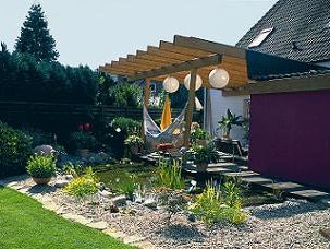 Schritt 12: Der Folienteich ist eine lebendige Wasserlandschaft geworden. Teichbau Schritt 12: Der fertige Folienteich mit Pflanzen und feinen Kieselsteinen umrandet liegt direkt neben der Terrasse mit einer gemütlichen Hängematte.