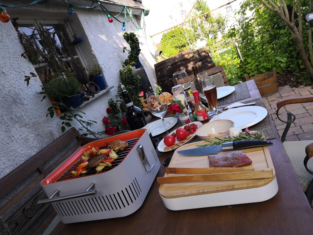 Keine Verbrennungsgefahr - Dank Hitzeschild steht der Cube direkt auf dem Holztisch! Gedeckter Gartentisch aus Holz, Fleisch, Messer, Grillzange und Rosmarin liegen auf dem Schneidebrett, der Everdure Cube steht daneben, auf dem Grillrost drei Gemüsespiese und zwei Rinderfilets
