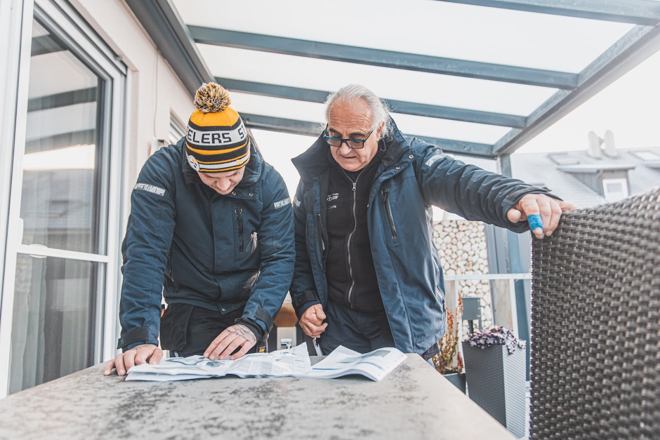 Zwei Monteure werfen auf überdachter Terrasse einen Blick in die Aufbauanleitung des Biohort Panorama mit Doppeltür