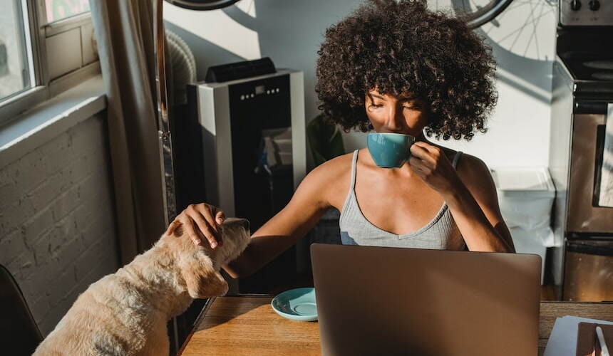 Junge Frau streichelt ihren Hund im Homeoffice Junge Frau mit dunklen Locken sitzt an einem Tisch mit Laptop, trinkt aus einer türkisfarbenen Tasse und streichelt einen Hund dabei