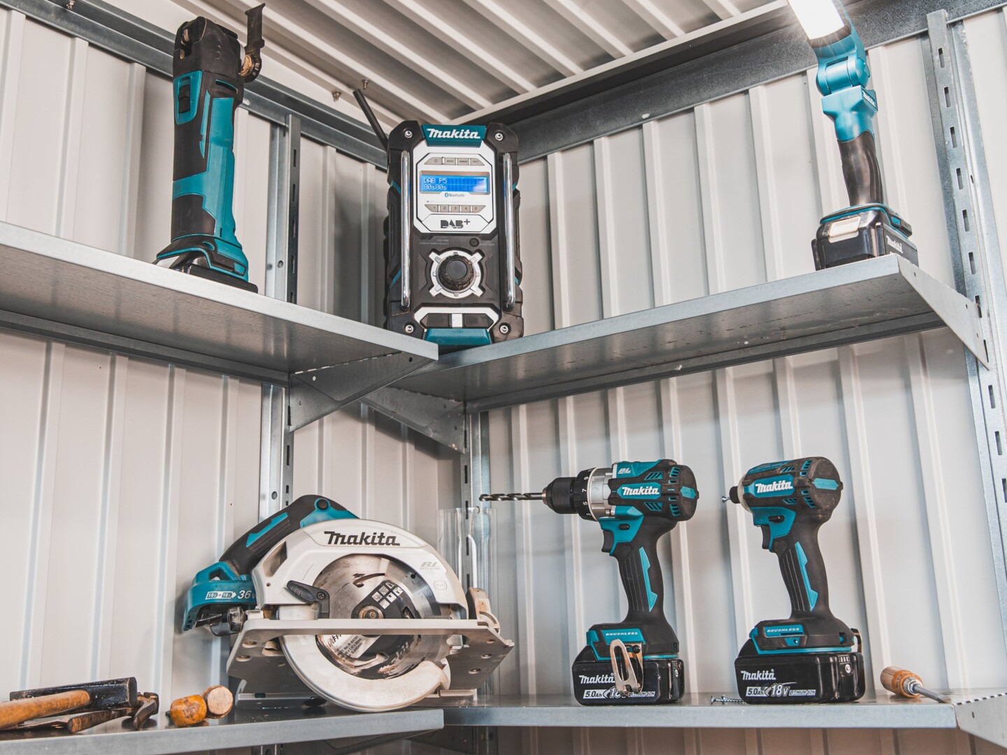 Werkzeuge der Marke Makita wie Akku-Handkreissäge, Akku-Bohrschrauber, Akku-Schlagschrauber, Akku-Baustellenradio und Akku-Arbeitsleuchte auf einem Wandregal im Gerätehaus.