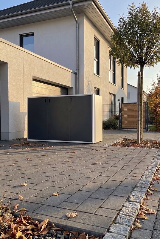 Eine große anthrazitfarbene Mülltonnenbox nimmt drei Tonnen neben der Garage auf.