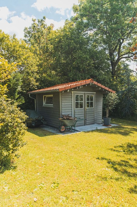 Graues Gartenhaus mit Ziegeldach und Schubkarre auf grüner Wiese